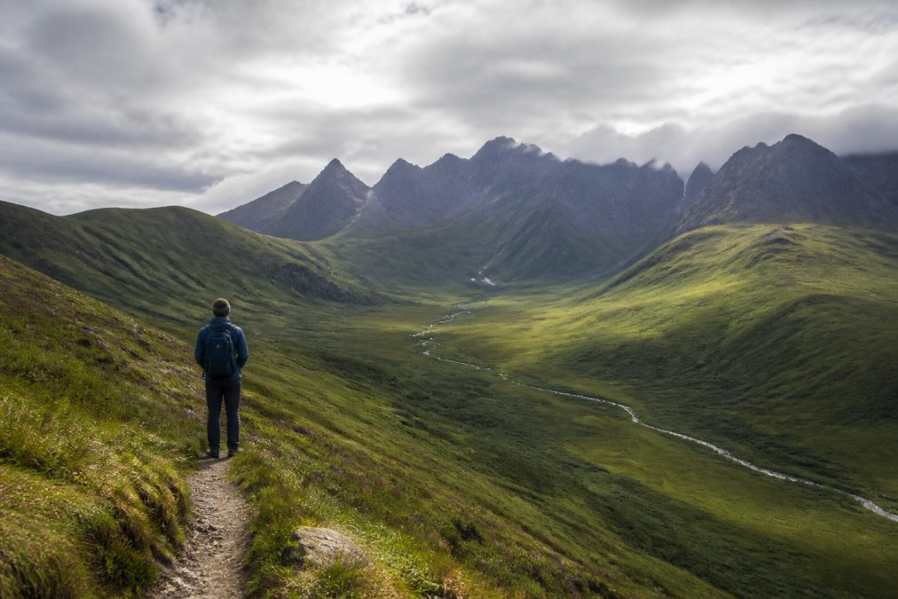 A lone walker with a small daypack standing on a grassy foothill footpath, looking across a green glen towards the jagged silhouette of the Black Cuillins under a wide Hebridean sky