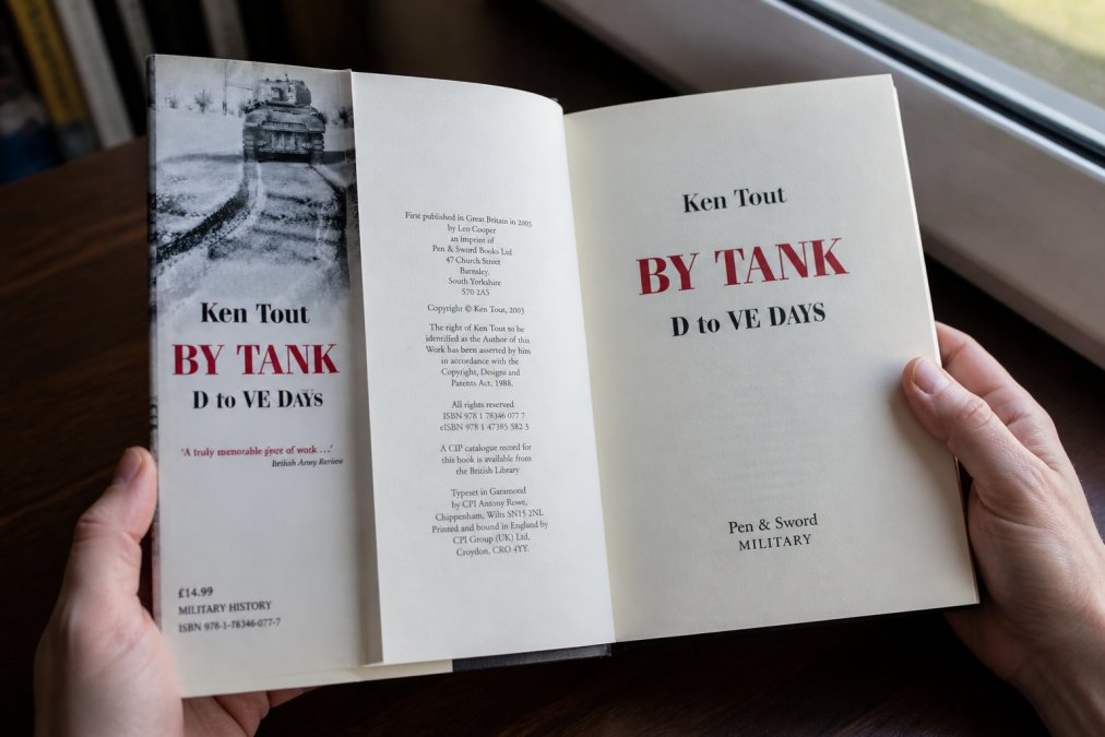 Close-up of hands holding an open hardcover military history book, showing the title page and copyright details with a dust jacket visible, natural daylight from a window illuminating the pages