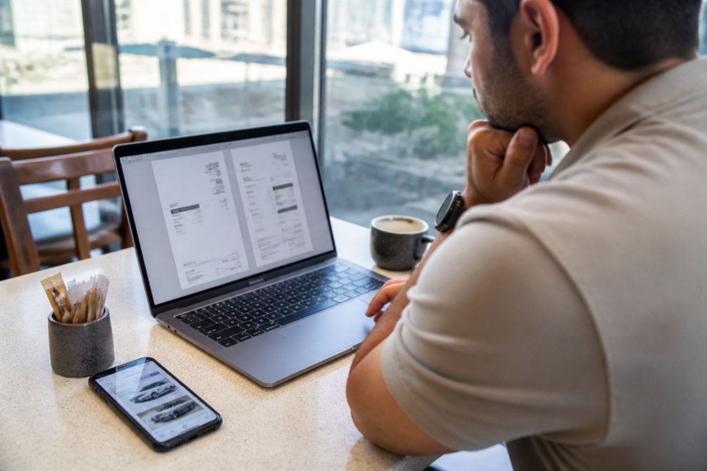 A professional in casual attire reviewing printed car repair estimates on a laptop at a bright Dubai cafe, with a smartphone displaying a garage comparison service beside the keyboard