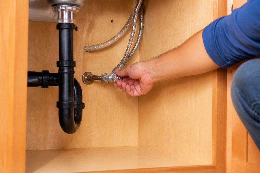 Homeowner shutting off water at the main valve under the sink with visible pipe connections and water shut-off handle clearly visible in bright natural lighting