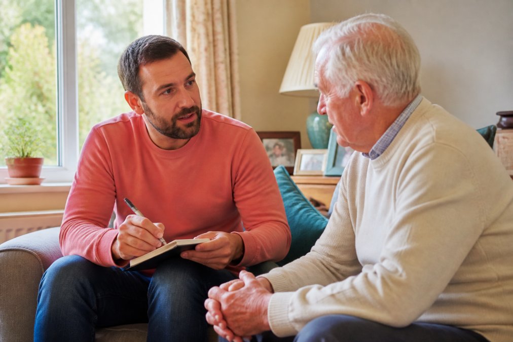 Adult son or daughter sitting with an elderly parent at home, having a thoughtful conversation with a notepad and pen, warm natural lighting from a window