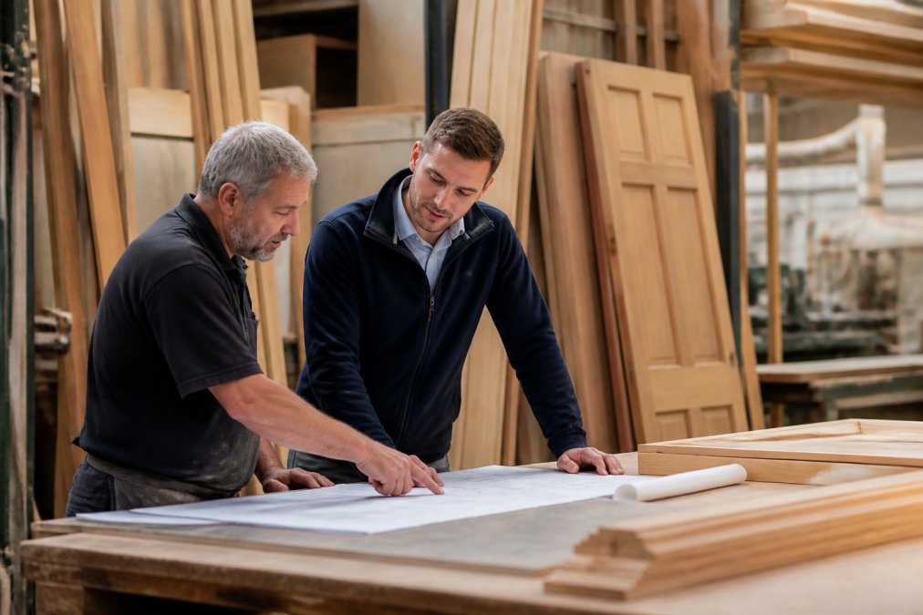 A craftsman in a UK joinery workshop reviewing technical drawings with a client, timber and finished doors visible in the background