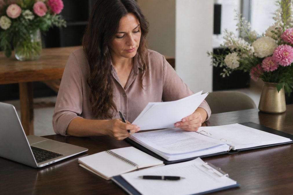 Person reviewing vendor documents and contracts at a desk with laptop, notepad, and event planning checklist visible, natural office lighting