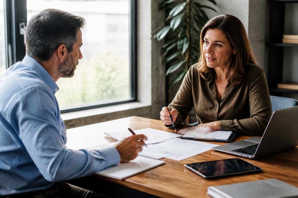 Business owner and consultant meeting in professional office setting, discussing strategy notes and documents across a wooden desk with laptop and tablet, natural window lighting