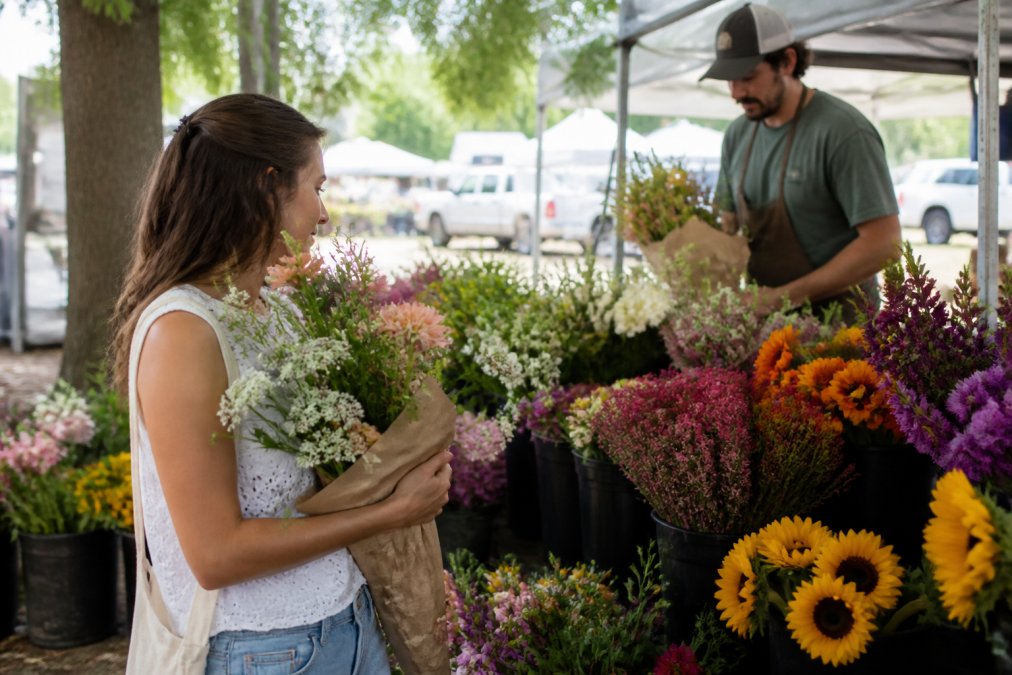 A woman browsing a vibrant farmers market flower stall, holding a bouquet of fresh seasonal blooms, with a farmer arranging buckets of flowers behind her