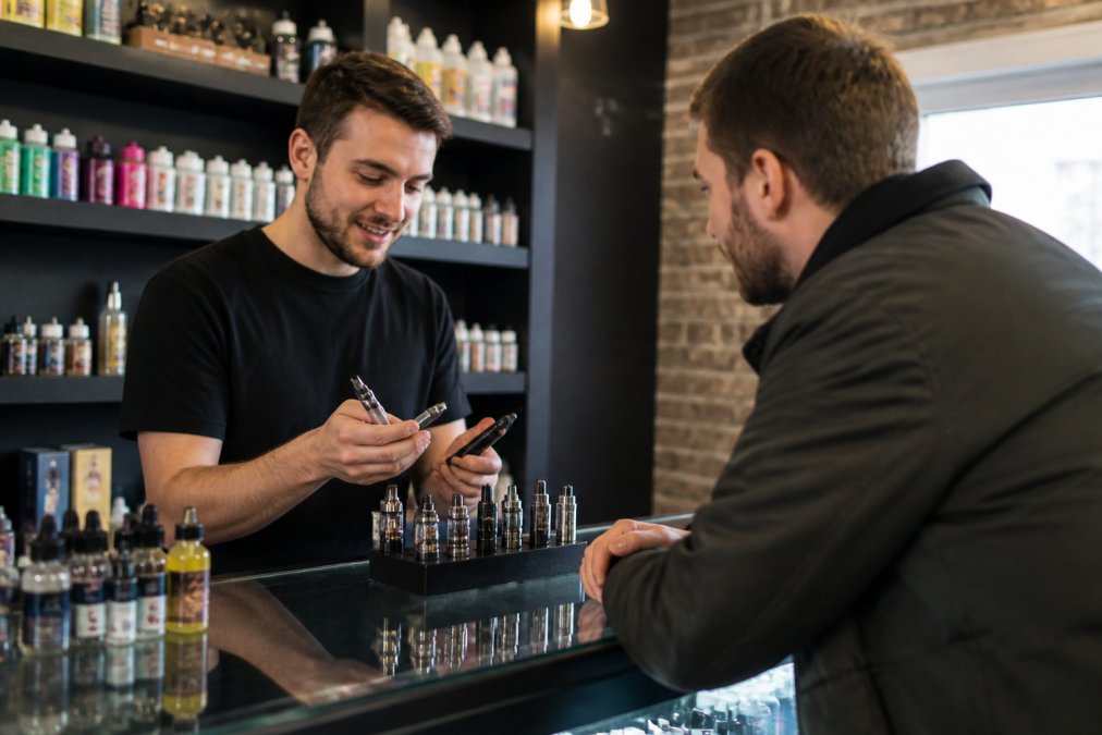 A friendly vape shop employee showing a customer different vape devices on a counter, with shelves of e-liquids in the background