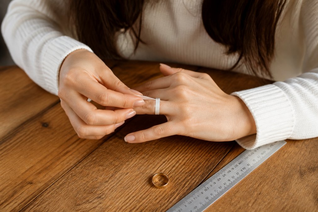 Close-up of a woman's hands wrapping a thin paper strip around her ring finger at a well-lit wooden table, with a ruler and a small ring resting nearby