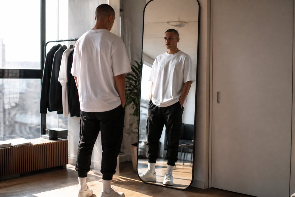 A young man wearing a heavy oversized white t-shirt paired with tapered black cargo joggers, standing in front of a full-length mirror in a modern apartment with warm natural window light