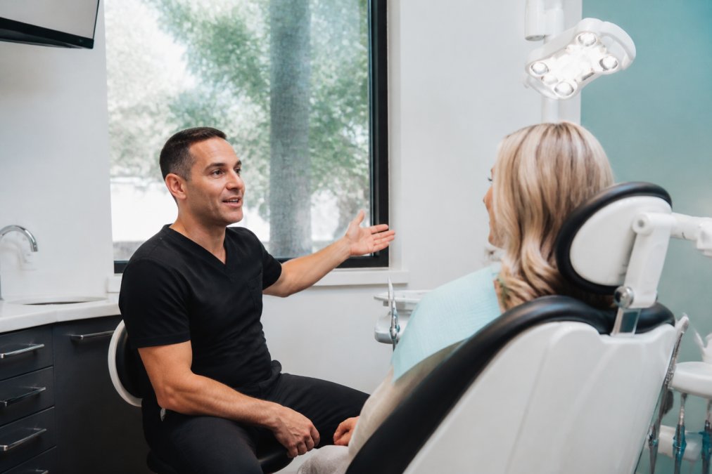 Dentist and patient in modern dental treatment room with patient seated in dental chair, dentist discussing digital treatment plan on wall-mounted monitor with warm clinical lighting