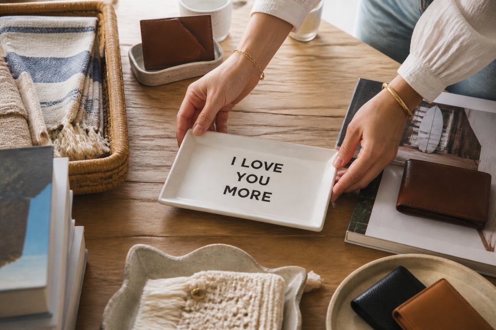 Hands carefully arranging curated gifts on a wooden table including ceramic trays, leather wallets, and lifestyle books, with soft natural light highlighting each item's texture and detail