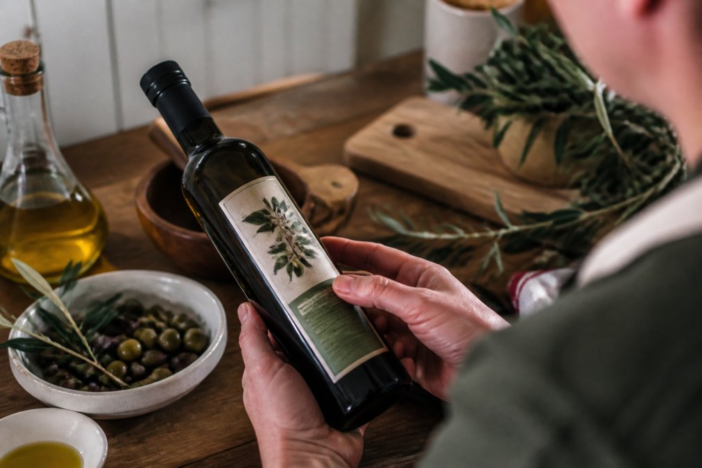 Close-up of hands holding a dark glass olive oil bottle, turning it to read the label with harvest date and origin details visible in warm kitchen lighting