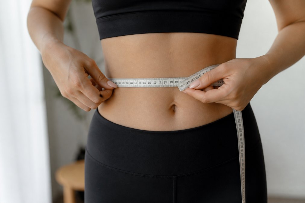 Close-up of a person using a soft measuring tape to measure waist circumference, tape positioned horizontally at the narrowest point of the torso, showing proper tension and body positioning