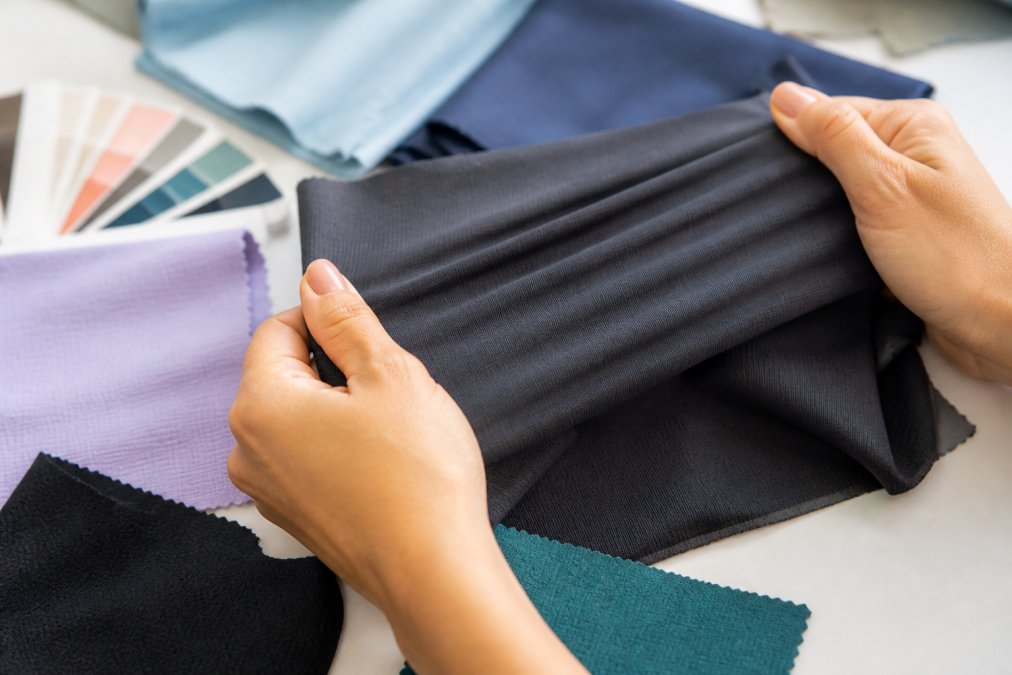 Hands stretching and testing different fabric samples showing varying degrees of elasticity and recovery, with close-up detail of the fabric weave texture and stretch response