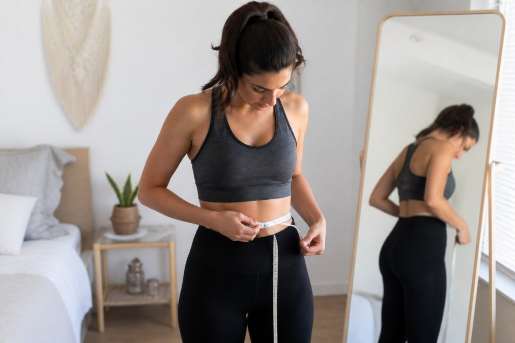 A woman in a bright bedroom standing in front of a mirror, holding a soft tape measure around her waist and looking down to read the measurement, wearing casual athleisure wear