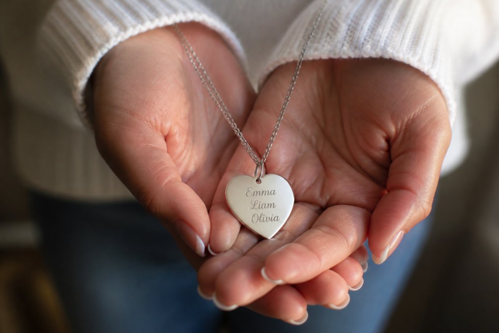 Close-up of a personalized heart name necklace being held in someone's hands, with soft natural lighting highlighting the polished stainless steel finish and delicate custom engraving
