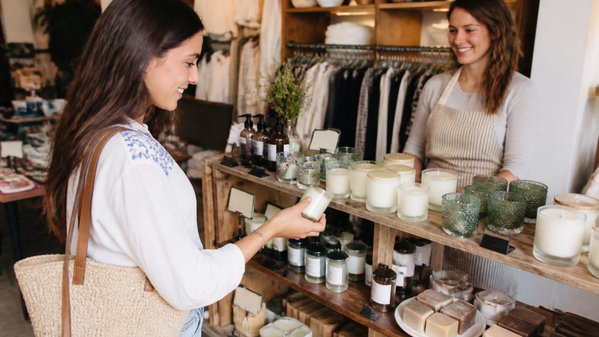 A smiling woman browsing a small boutique shelf filled with artisan candles, soaps, and home decor items, with a shop owner nearby