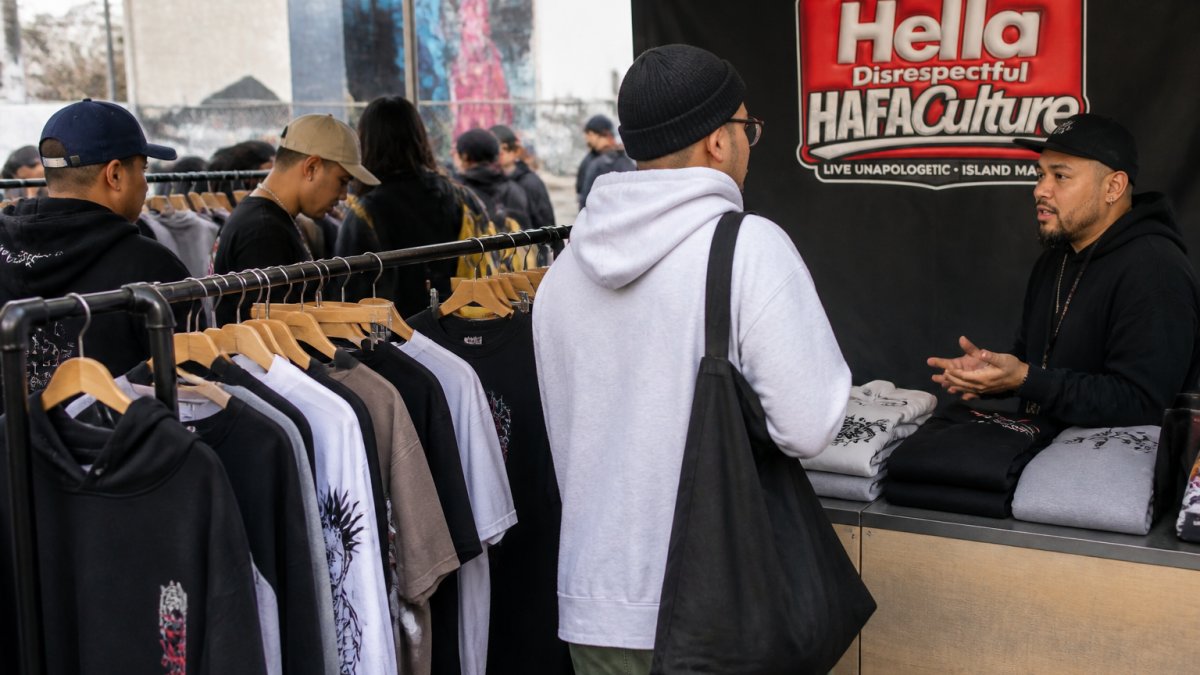 Shoppers browsing racks of graphic tees and hoodies at an independent streetwear pop-up market, with a vendor talking to a customer in the foreground
