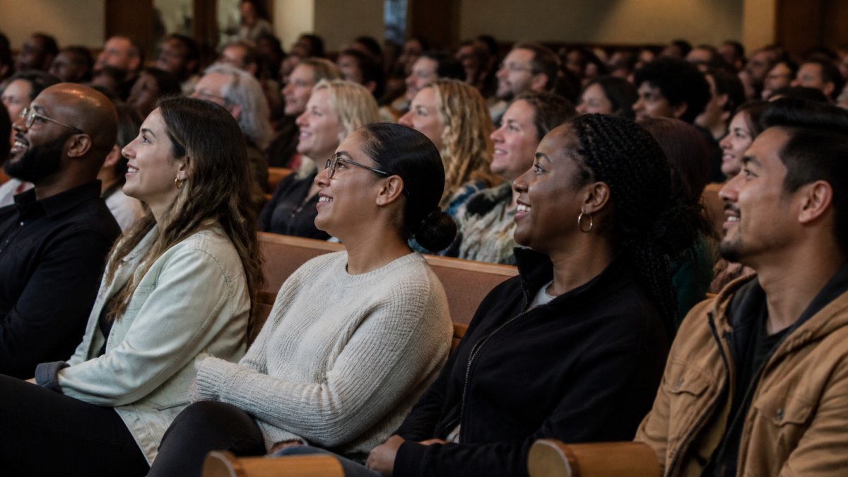 A diverse group of people sitting together in a church sanctuary during worship, smiling and engaged, with warm lighting and an inclusive atmosphere