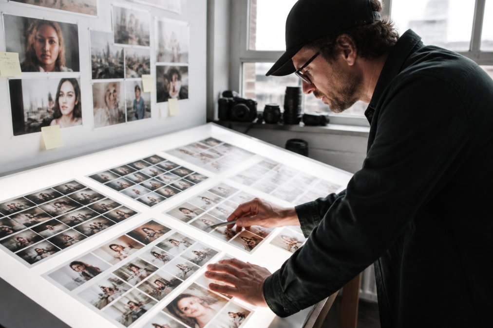 A photography director reviewing a large printed contact sheet on a light table, comparing competitor images and brand mood board photos side by side in a sunlit studio