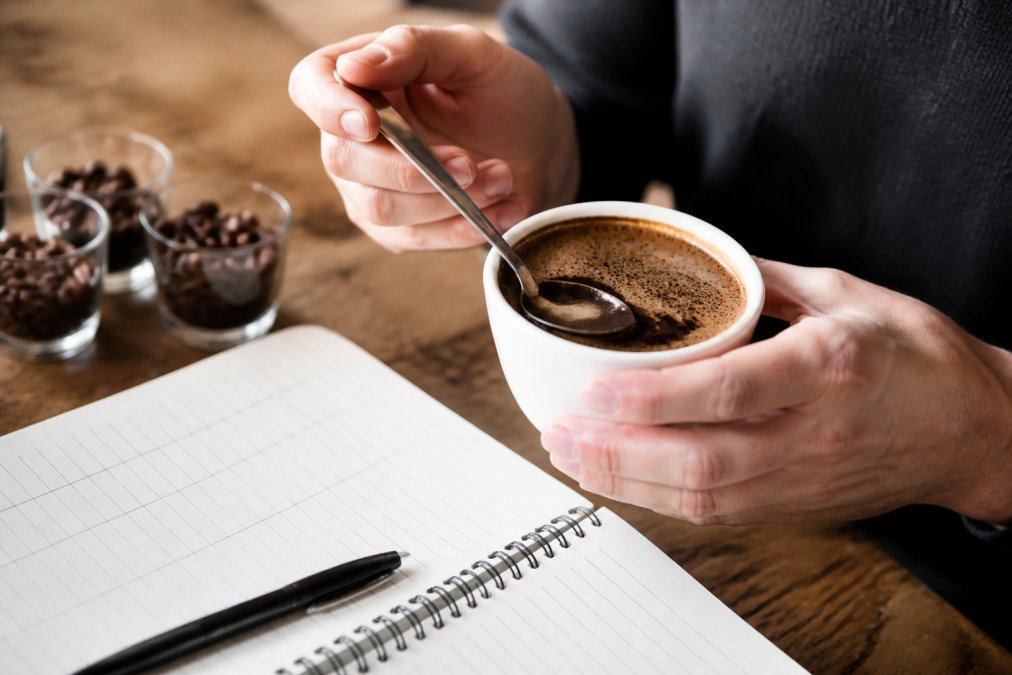 Close-up of hands holding a white cupping bowl filled with brewed coffee, with a cupping spoon resting in the bowl and a notebook with tasting notes visible on a wooden table