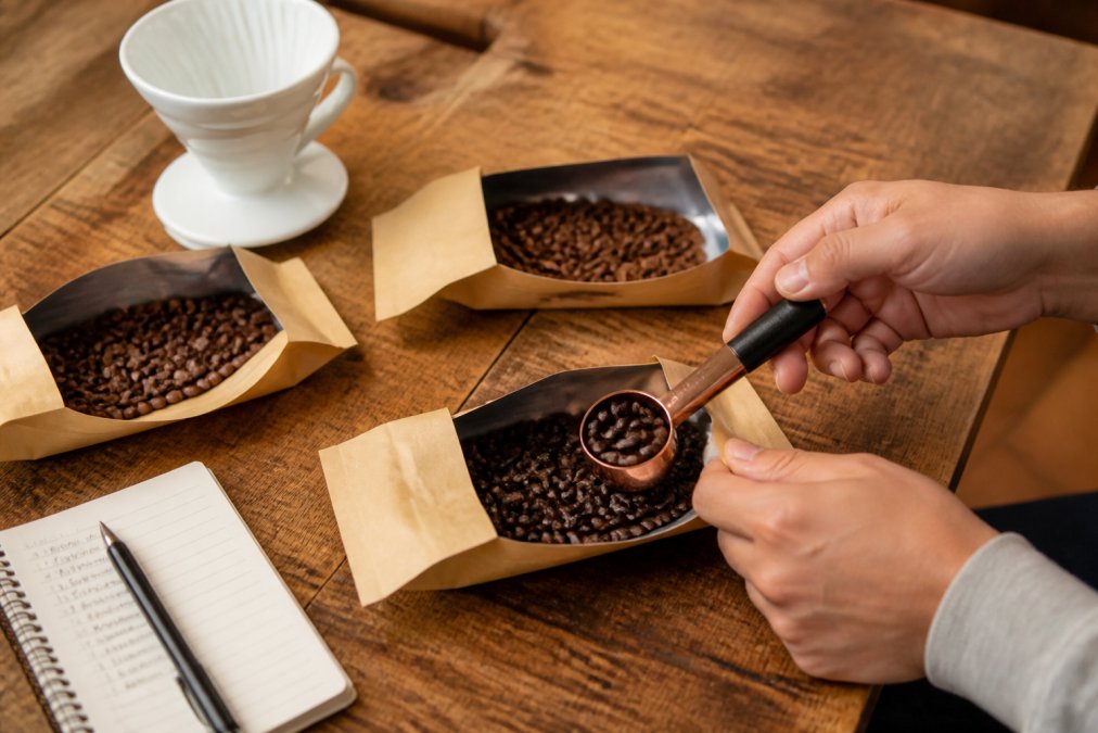 A wooden table displaying three open Coffee Depot sample packs with different roasted beans visible, a white ceramic brewing vessel, a small notebook with tasting notes, and hands holding a coffee scoop over one of the packs