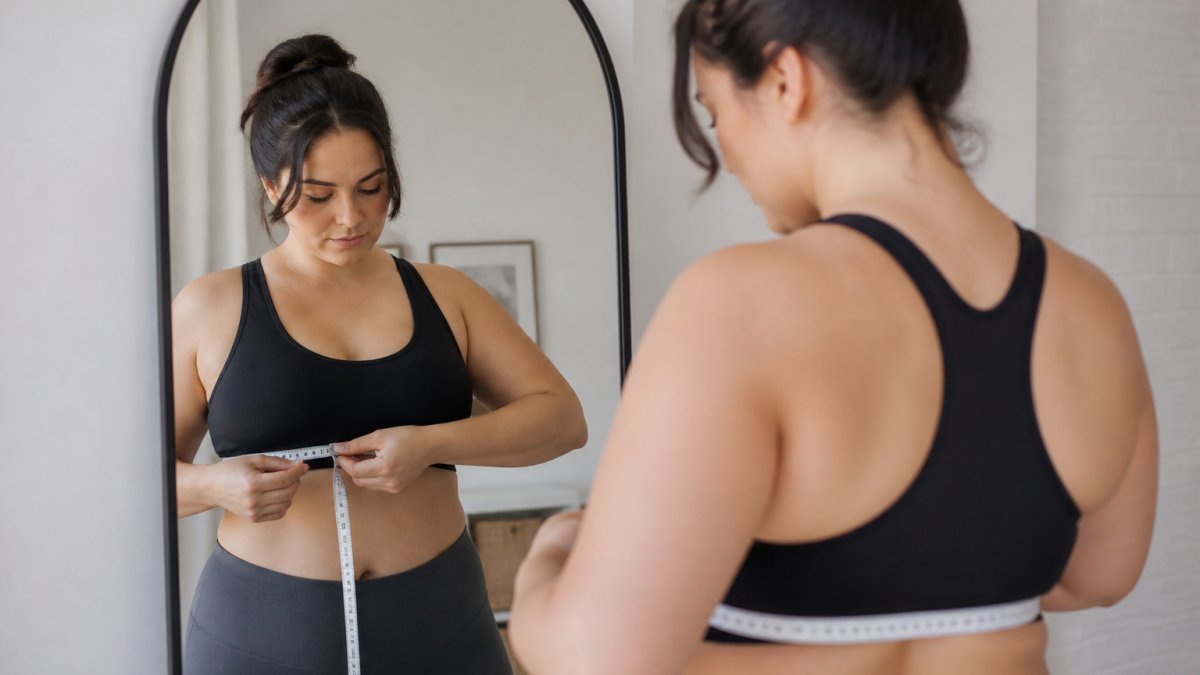 Woman standing in front of a mirror with a measuring tape wrapped snugly around her underbust, showing proper measuring posture and tape placement