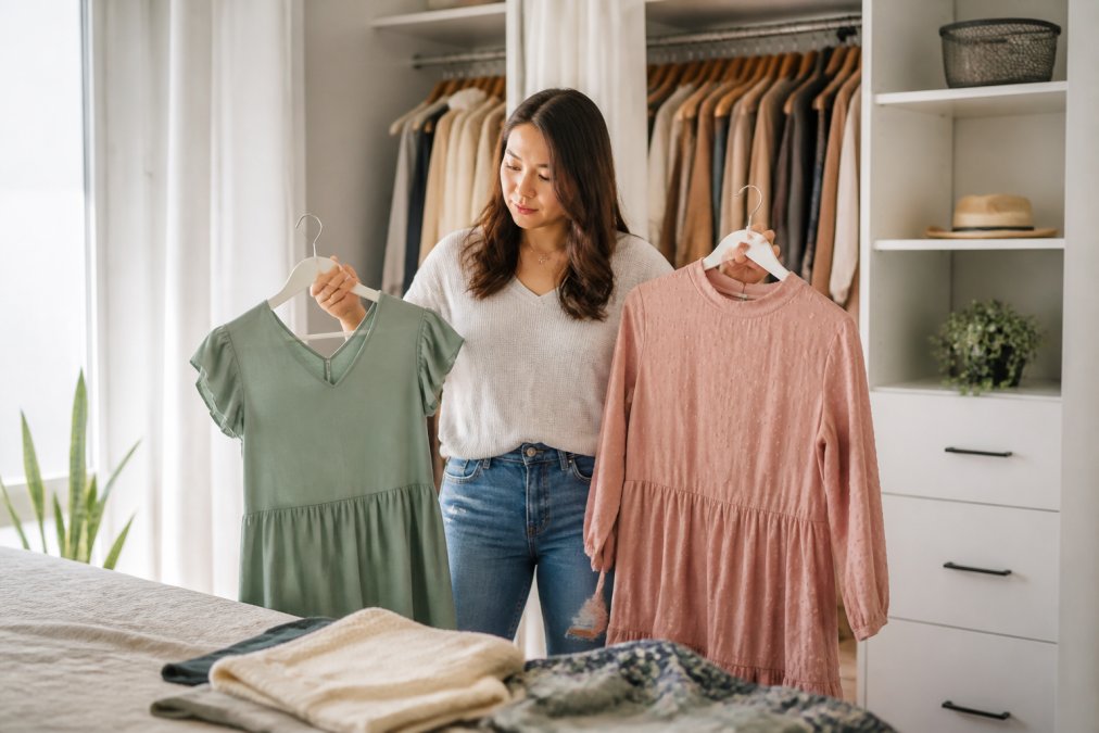 A woman standing in front of an open, organized closet, holding two different outfits on hangers and comparing them thoughtfully, natural light, relaxed home setting