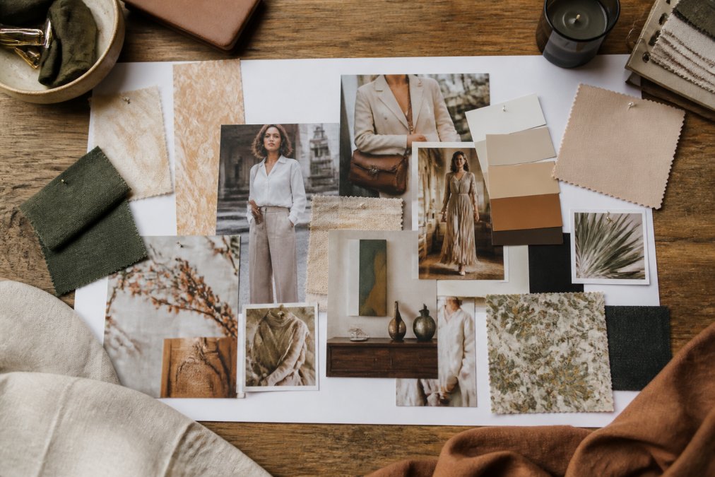 A flat lay of a physical mood board on a wooden table with magazine clippings, fabric swatches, printed photos, and color paint chips arranged in an artistic collage