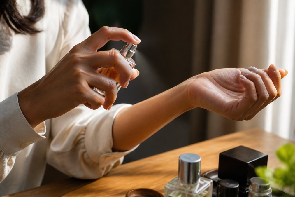 Close-up of a person's inner wrist held up to a small glass perfume bottle, warm natural afternoon light, the skin slightly flushed from testing fragrance, suggesting careful evaluation of scent on bare skin