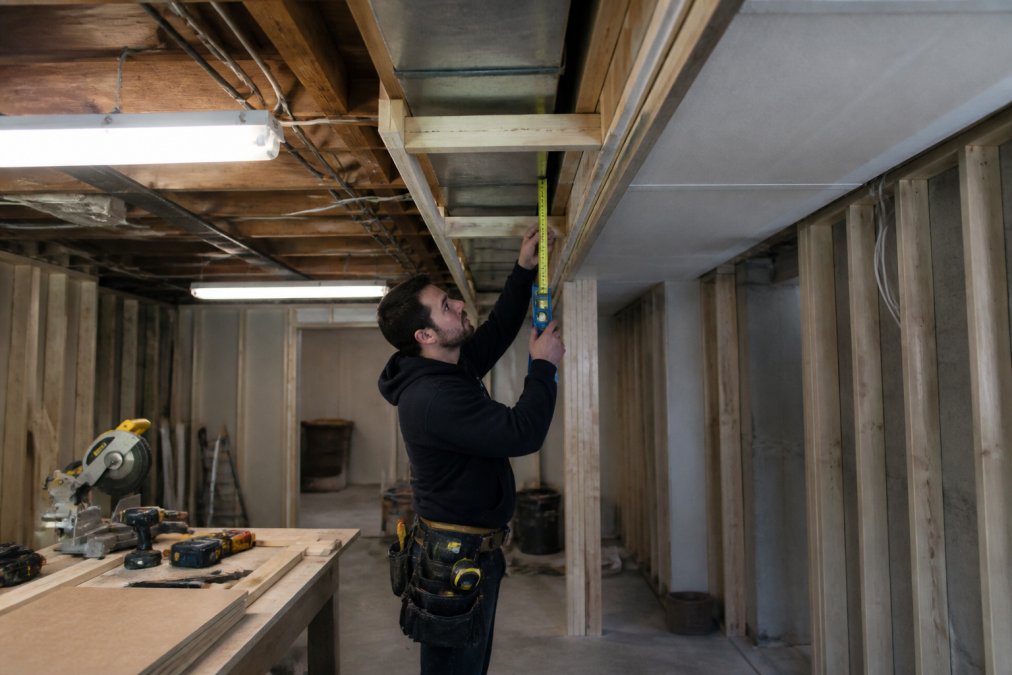 Professional HVAC contractor installing framing around rectangular ductwork in basement, measuring clearance between soffit frame and duct bottom with level and measuring tape visible, fluorescent work lights illuminating the space