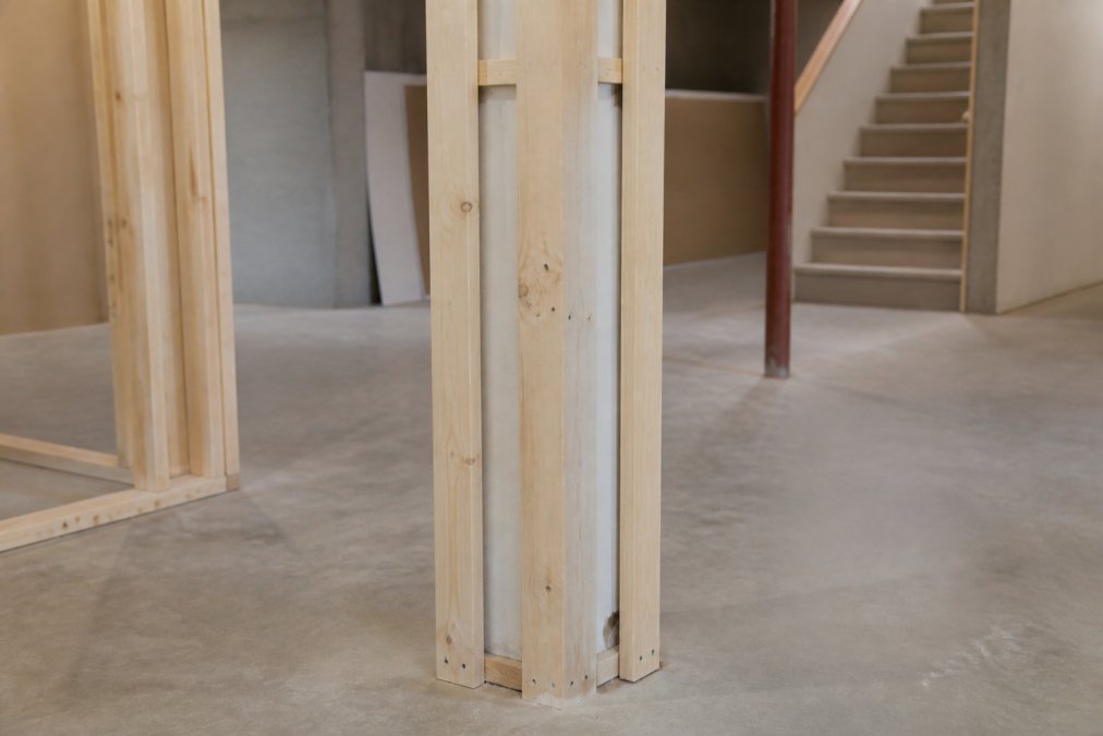 Close-up of a basement support post wrapped with furring strips and shims, showing the framing structure before drywall installation