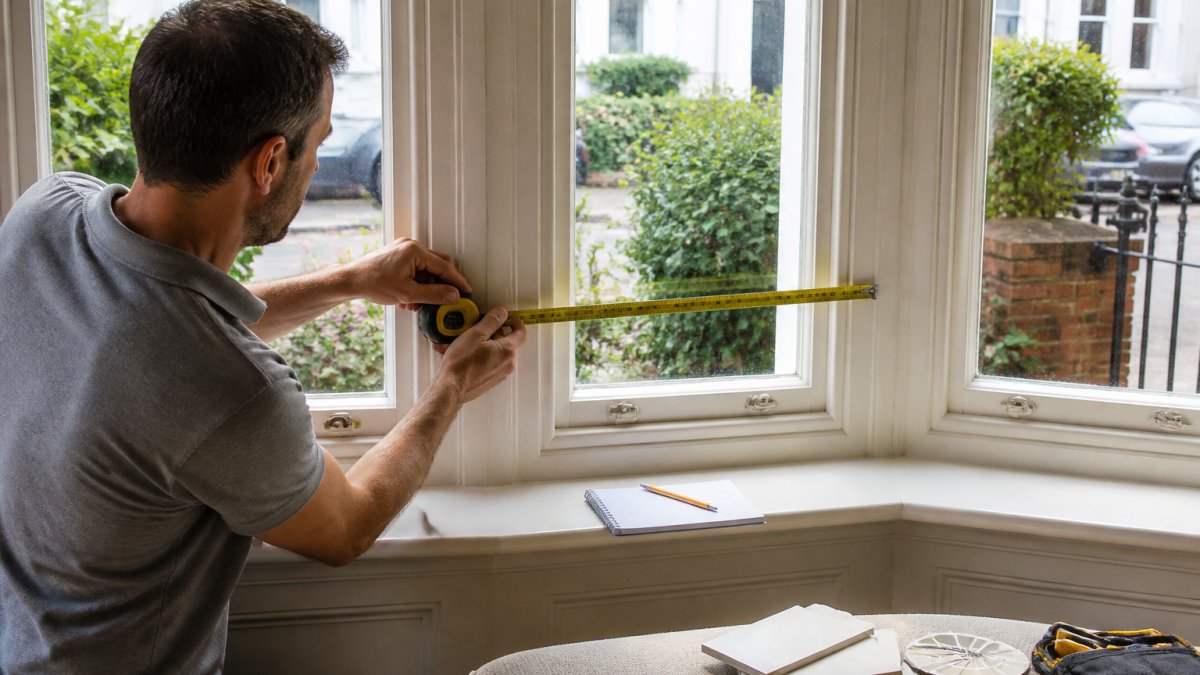 A person using a metal tape measure across the angled panel of a curved bay window in a period British home, with a notepad and pencil on the windowsill nearby
