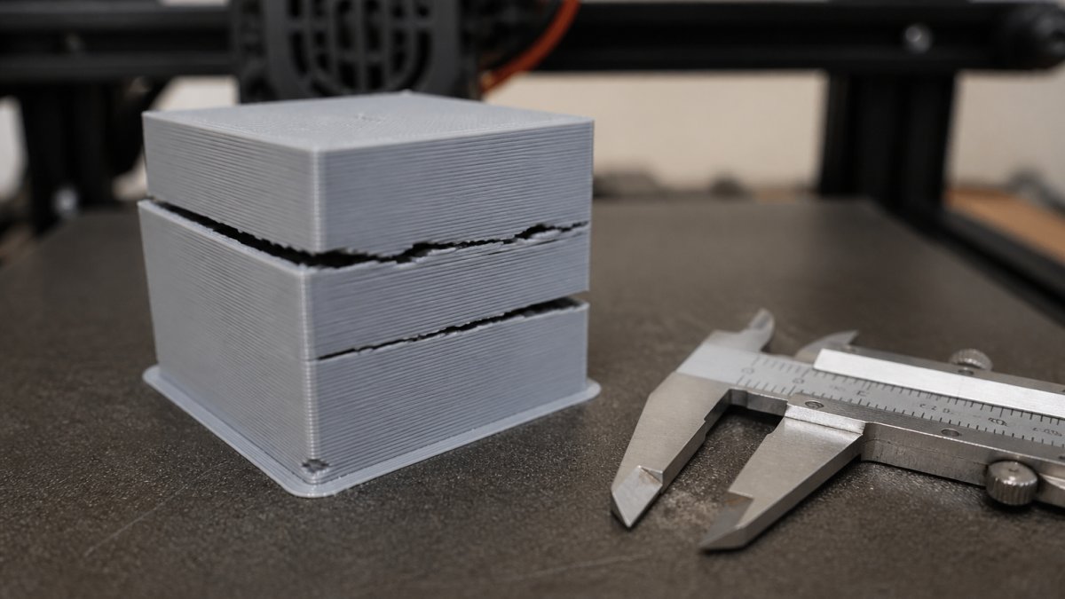 A close-up photograph of a 3D printed object on a printer bed, showing visible layer shifting and gaps, with a pair of calipers resting beside it for scale