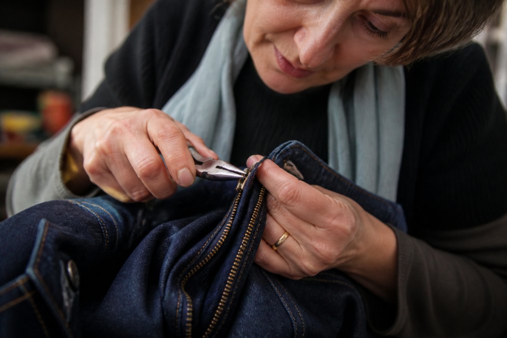 Close-up of hands holding needle-nose pliers gently adjusting a metal zipper slider on a denim jacket, with focused lighting on the repair work