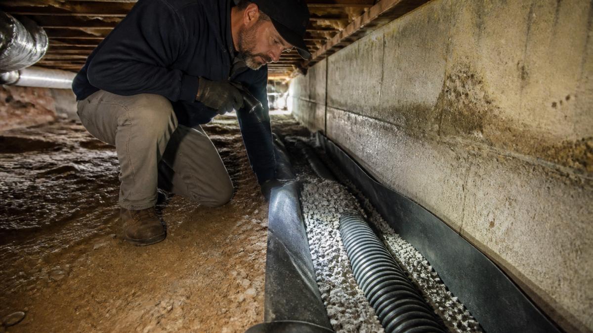 A homeowner in work clothes kneeling in a shallow dirt crawl space, inspecting a French drain trench lined with gravel along the foundation footing, with a flashlight illuminating the area