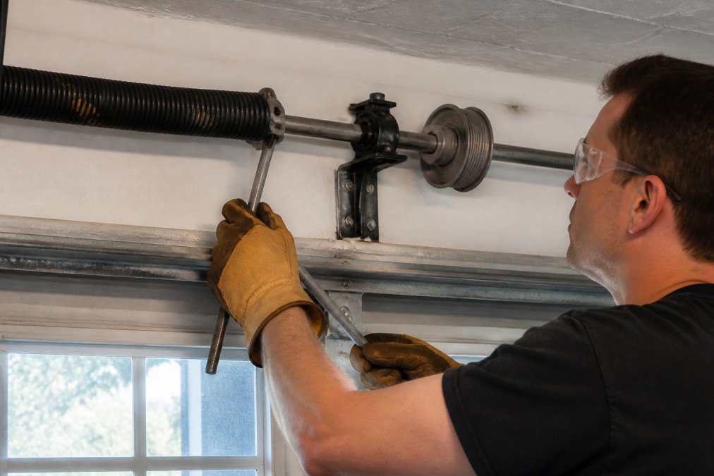 A technician in safety glasses and leather gloves carefully inserting a steel winding bar into the winding cone of a torsion spring mounted above a garage door, with the cable drum and center bearing bracket clearly visible in the background under overhead garage lighting