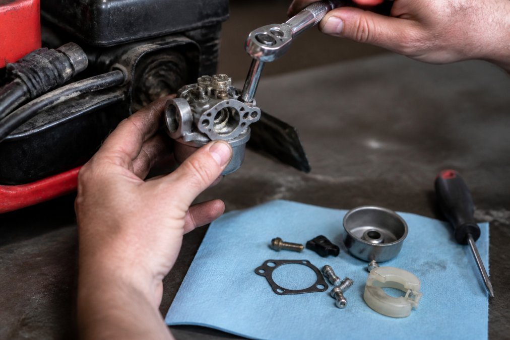 Hands using a socket wrench to remove a lawnmower carburetor from an engine on a workbench