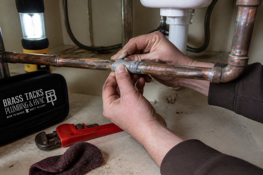 Close-up of a homeowner's hands pressing epoxy putty onto a corroded copper pipe under a kitchen sink, with a pipe wrench and abrasive cloth resting on the cabinet floor nearby, illuminated by a work light