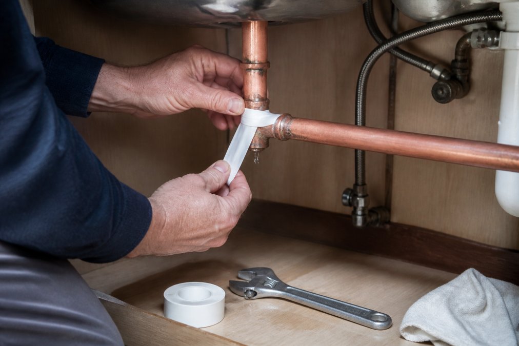 Hands wrapping silicone tape around a leaking copper pipe joint to fix a leaking pipe under a kitchen sink.