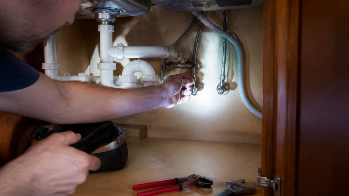 A homeowner's hand turning the shutoff valve under a kitchen sink, with a flashlight illuminating the pipes and cabinet interior