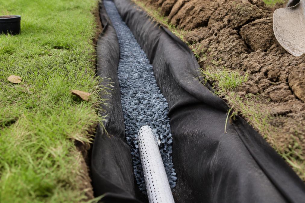 A close-up of a shallow trench cut into a lawn, lined with landscape fabric and partially filled with blue metal gravel, with a perforated pipe visible
