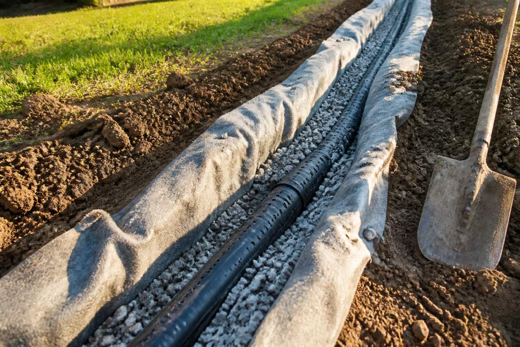 A close-up of a partially installed French drain in a backyard trench, showing the perforated pipe surrounded by gravel and wrapped in filter fabric, with a shovel resting nearby and late afternoon sunlight casting long shadows
