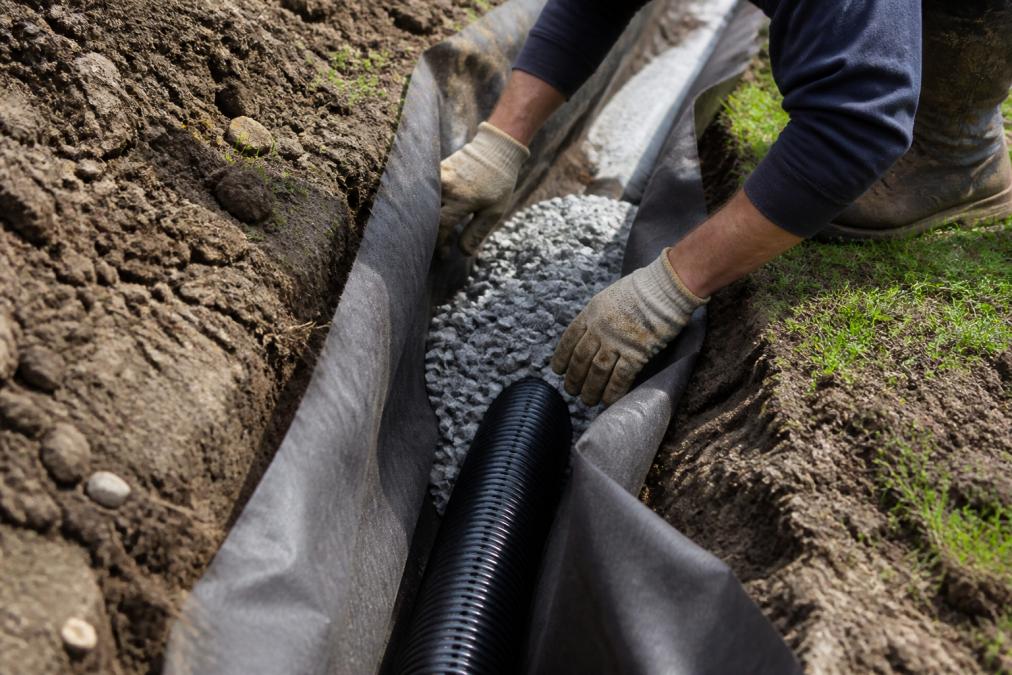 Close-up of a trench being excavated in a residential backyard, showing layers of landscape fabric, perforated drainage pipe, and washed stone beginning to be placed, with a contractor's hands visible positioning materials