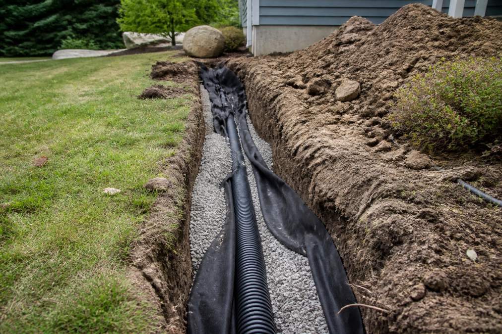 A contractor installing a French drain in a residential backyard, laying perforated pipe wrapped in filter fabric inside a gravel-lined trench on a sunny afternoon