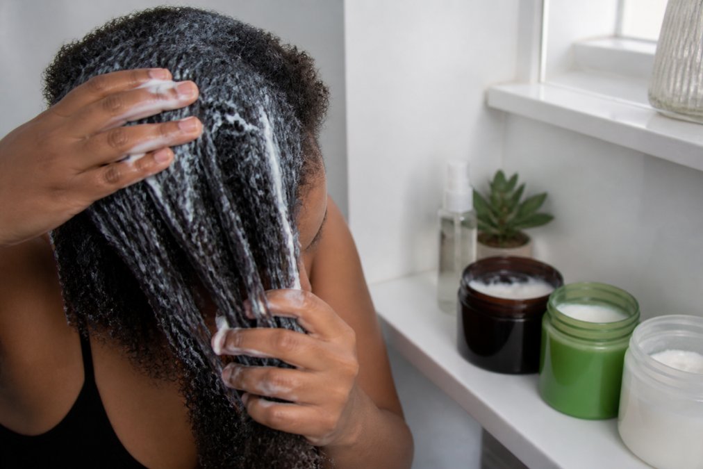 Close-up of hands applying a thick, white deep conditioning hair mask to damp, textured natural hair, with several open product jars arranged on a white bathroom shelf in soft natural light