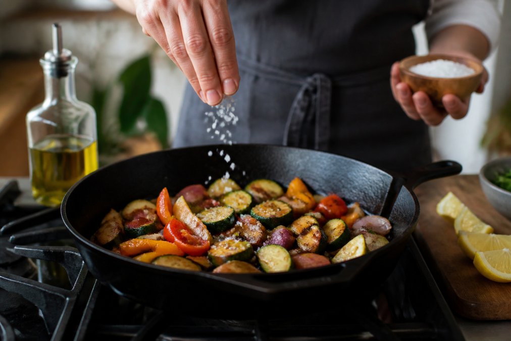 A home cook's hands sprinkling flaky sea salt over a colorful sautéed vegetable dish in a cast-iron skillet on a stove, with lemon wedges and a bottle of olive oil nearby on a wooden countertop