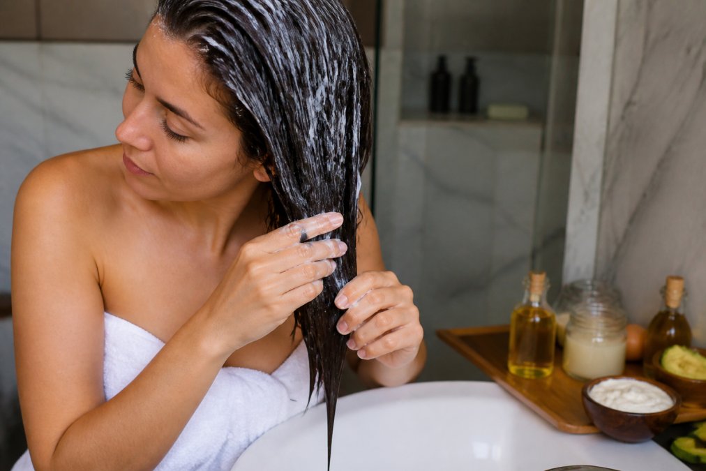 Close-up of a woman applying a creamy white hair mask to her damp hair over a bathroom sink, with natural oil bottles and a wooden bowl of ingredients visible on the counter, warm bathroom lighting