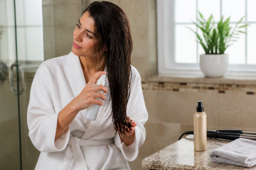 Woman applying leave-in conditioner spray to the ends of her towel-dried hair in a bright bathroom, with a heat protectant bottle and microfiber towel visible on the vanity counter