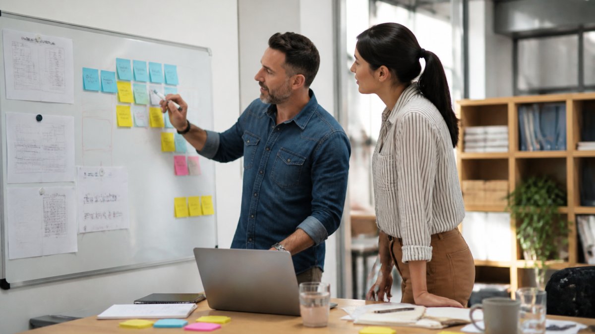 A small business owner and a team member reviewing workflow notes on a whiteboard in a bright office, with sticky notes showing process steps and a laptop open on the table