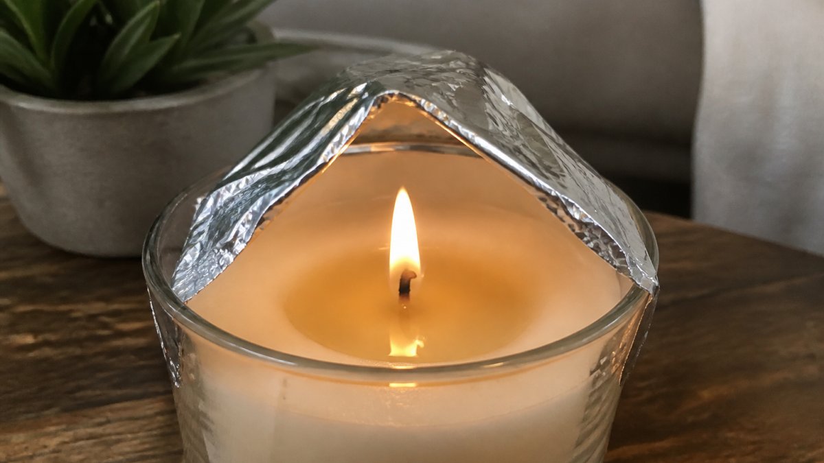 Close-up of a lit candle with aluminum foil tent positioned around the wick, showing the melt pool beginning to widen as heat reflects inward